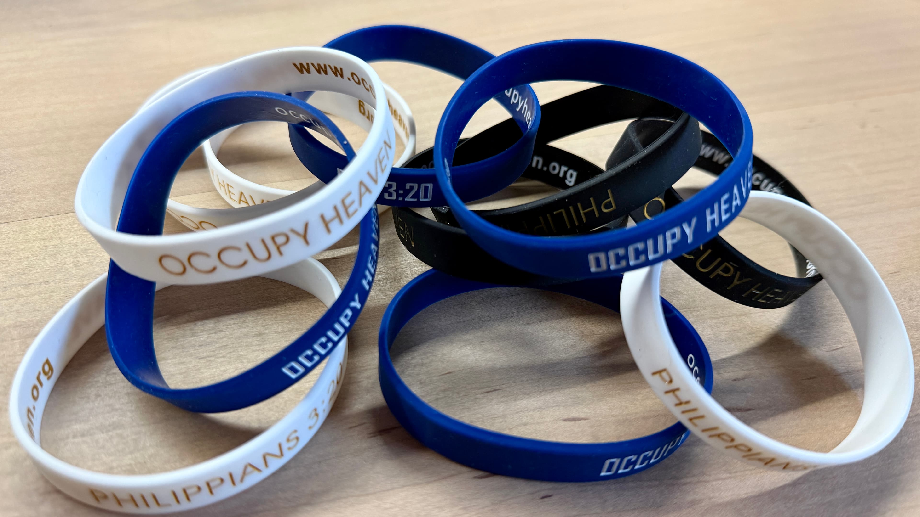 A pile of Occupy Heaven bracelets in blue, white, and black, engraved with Philippians 3:20 and OccupyHeaven.org